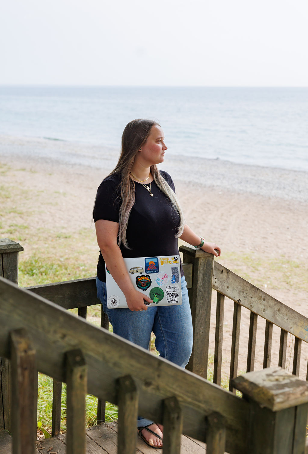 Dana stands on stairs overlooking the beach with her laptop, contemplating when to hire a bookkeeper for a small business.