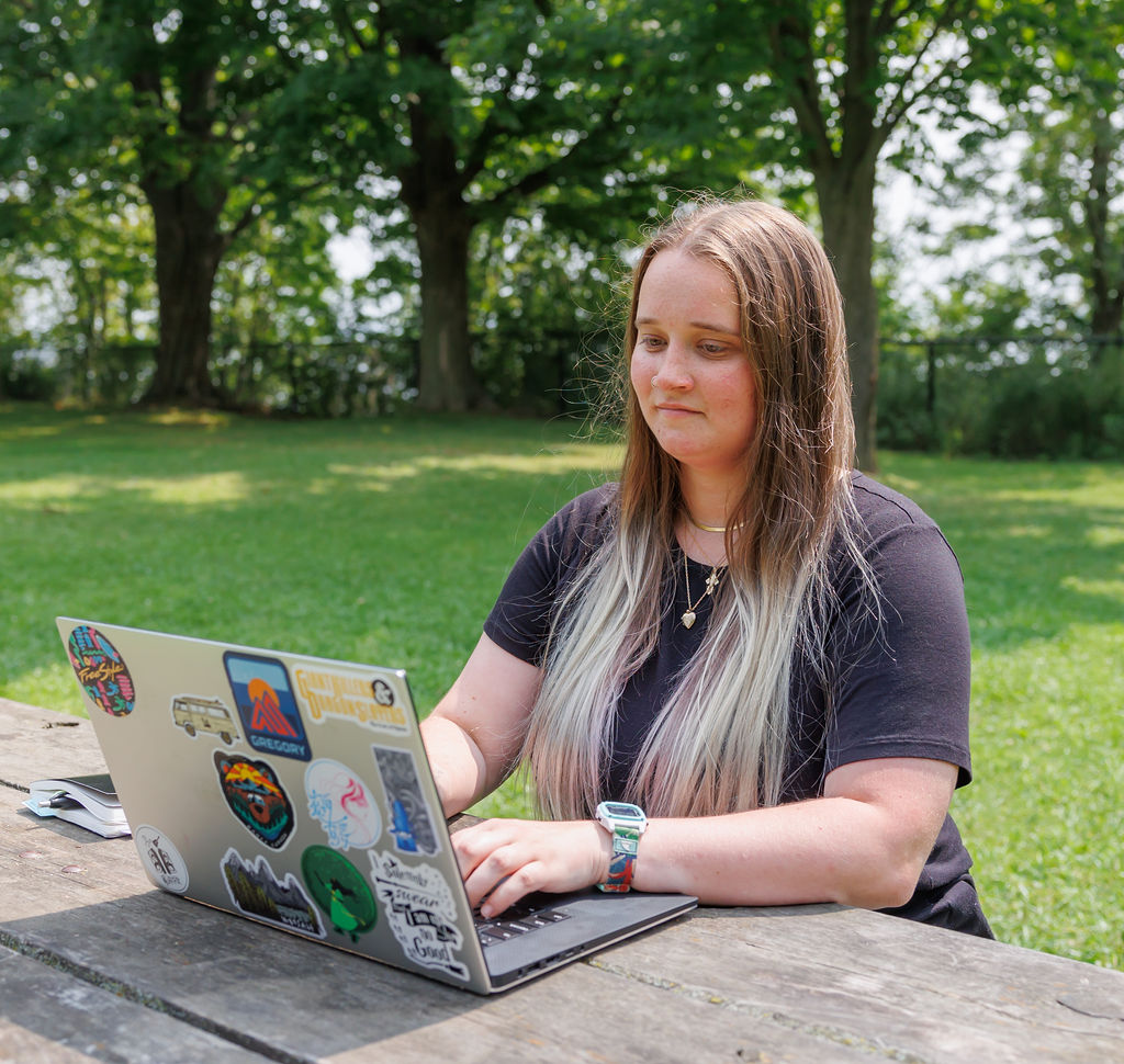 Dana, sitting at a picnic table outside, writing a blog post on a laptop about why Wealthsimple is the best bank for Canadian small business owners.